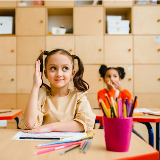 Female student raising her hand while sitting at a classroom desk, and one other students behind her is paying attention to another person or object off-camera.