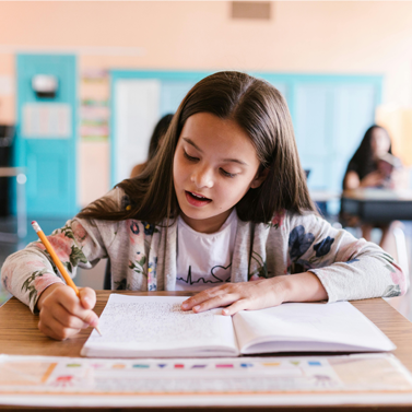 Pre-adolescent girl sitting at a desk inside a classroom and writing with a pencil.