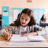 Pre-adolescent girl sitting at a desk inside a classroom and writing with a pencil.
