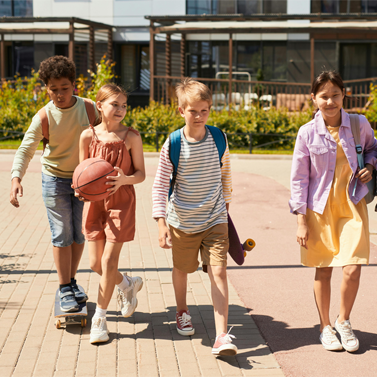 Four kids walking through a park, with one riding a skateboard, one holding a skateboard, while another is holding a basketball.