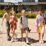 Four kids walking through a park, with one riding a skateboard, one holding a skateboard, while another is holding a basketball.