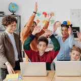 Two students sitting in front of laptops while students behind them cheer them on, and an adult standing on the left side.