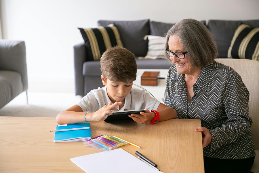 A younger child using a tablet in front of a smiling older woman who is sitting at a table next to the child.