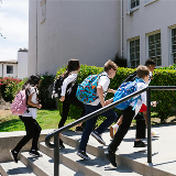 Side view of five middle school-aged children running up a set of stairs that lead into a school