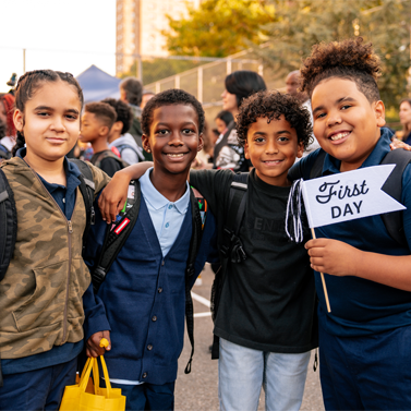 Four kids in school yard smiling with each other, and one student holding up a flag that says, "First Day."
