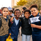 Four kids in school yard smiling with each other, and one student holding up a flag that says, "First Day."