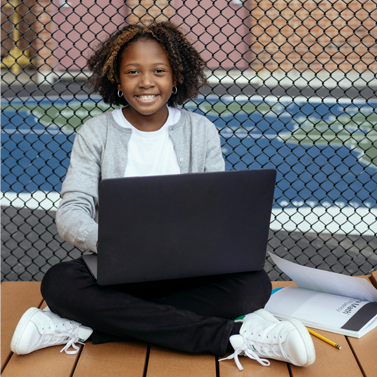 Middle school-aged girl sitting on a park bench with an open laptop and a nearby notebook