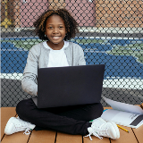 Middle school-aged girl sitting on a park bench with an open laptop and a nearby notebook
