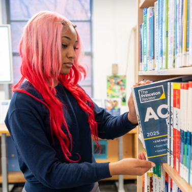 A teenage girl with pink and red hair pulls a copy of The Princeton Review off of a shelf in a library.
