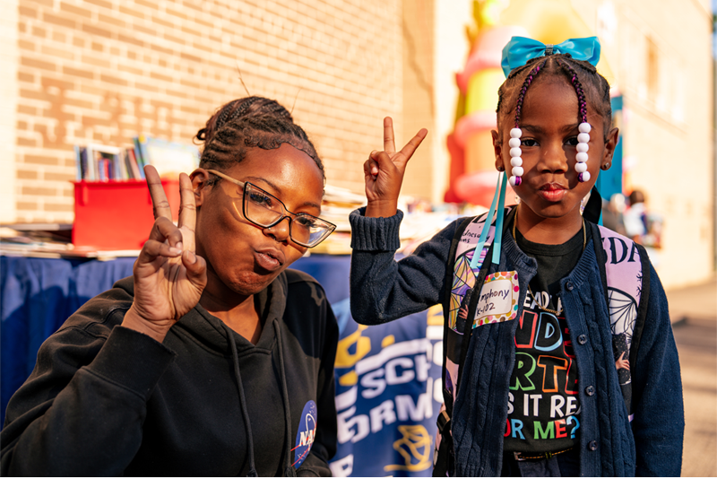 Close up of a mother (left) and her daughter (right) holding two-fingers in 'peace' sign.