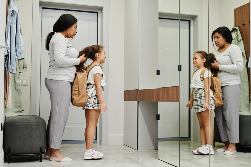 Side view of an older woman, a mother, standing behind a younger girl, her daughter, and holding the girl's hair up in her hands like she's getting ready to style it into some kind of ponytail or bun.