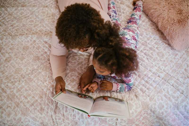 Top view of a mother lying face down on a bed with her daughter while reading a book together.