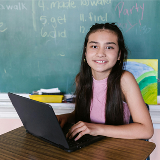 A middle schooler with a pink shirt sitting at a desk with a laptop inside of a classroom.