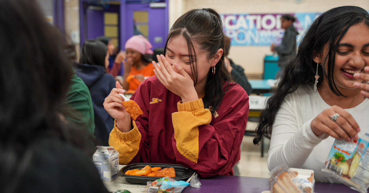 Two students sitting in a school cafeteria laughing while one student picks up food from her plate.