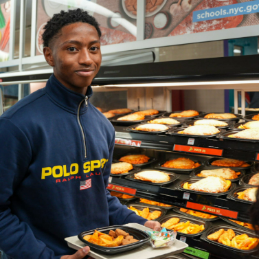 A student holding a lunch tray in a school cafeteria smiles in from of an array of school meals.