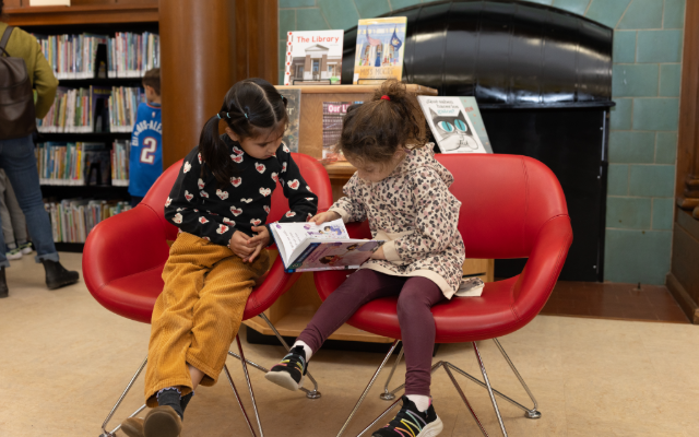 Two girls sitting in red chairs reading a book together in a library.