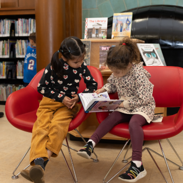 Two girls sitting in red chairs reading a book together in a library.