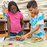 Two children standing at a table in front of a bookcase at the library.