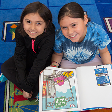 Two girls sitting on a blue carpet. The girl on the right is holding an open book in her lap. The girls are looking up and smiling at the camera.