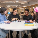 Four students sitting at a table with a stack of books on it. The second student from the left has an open book in front of her that they are looking at together.