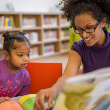 A woman is reading a picture book aloud to a young child in a library.