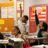 Students at desks in a classroom with many posters. Two students in the back are excitedly raising their hands, while a student in front of them is looking at a book.