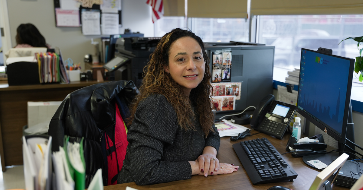 Photo featuring a female parent coordinator smiling at the camera while sitting at a school office computer desk.