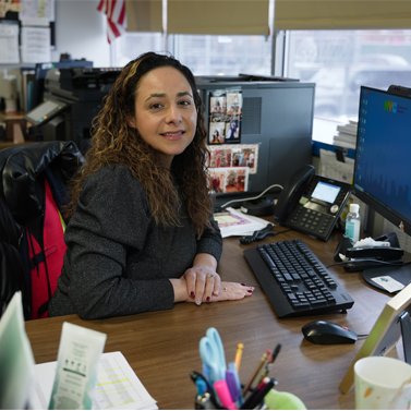 Photo featuring a female parent coordinator smiling at the camera while sitting at a school office computer desk.