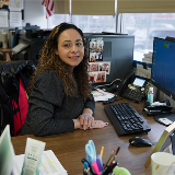 Photo featuring a female parent coordinator smiling at the camera while sitting at a school office computer desk.