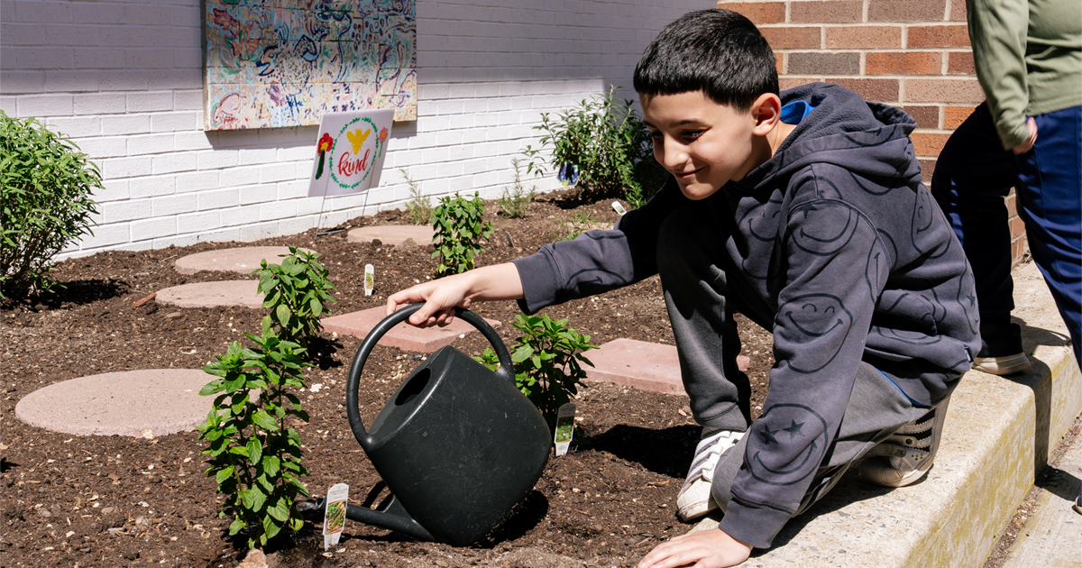 A young middle school-aged student sitting by a school garden and holding a watering can.