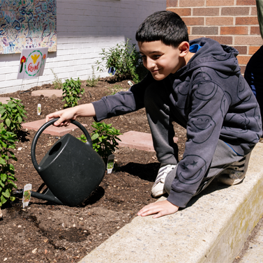 A young middle school-aged student sitting by a school garden and holding a watering can.