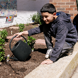 A young middle school-aged student sitting by a school garden and holding a watering can.