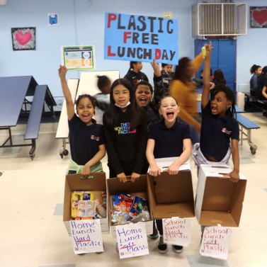 Five students at P.S. 188 The Island School in Manhattan hold up a sign with Plastic Free Lunch Day written on it, and display the plastic waste they have collected from their cafeteria, which is sorted into four cardboard boxes.