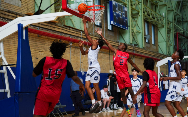 Boys playing basketball. A boy in a red uniform is reaching to block a boy in a white uniform from making a basket. The basketball is near the hoop.