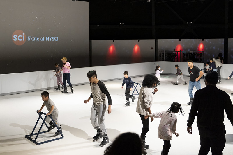 Skating rink featuring several groups of families skating in front of a projector screen that reads, 