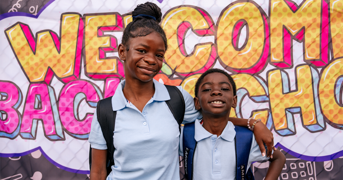 Sister and brother standing next to each other in front of a mural that reads, "Welcome Back to School"