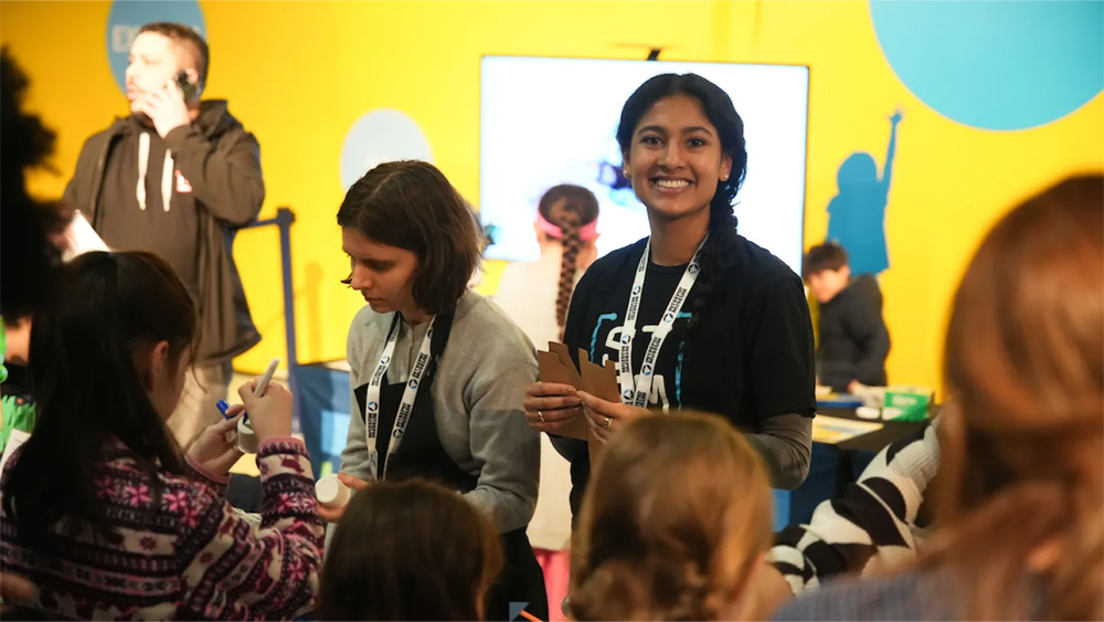 Two young women standing in front of a class of younger students, with one looking directly at the camera and smiling.