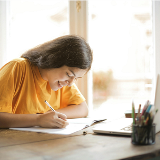 Side view of a young woman in a yellow shirt writing in a notebook while sitting at a table with a laptop on it.