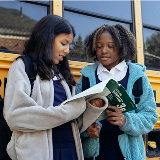 Two middle school-aged girls reading a grammar workbook together while standing on the side of a yellow bus.