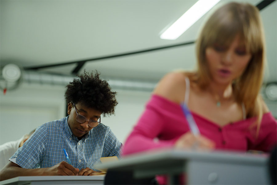 Two students in a classroom, sitting at their desks and writing in their notebooks.