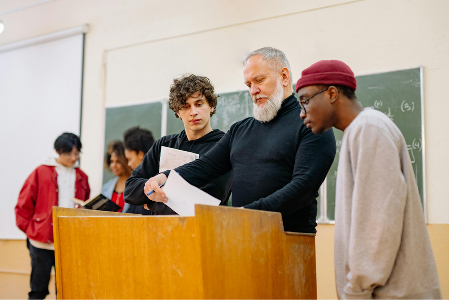 Two students alongside a male teacher who is overlooking a paper while standing at a lecturn.
