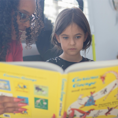 Close-up of a young child reading a picture book while a teacher happily looks on from the left side.