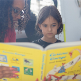 Close-up of a young child reading a picture book while a teacher happily looks on from the left side.