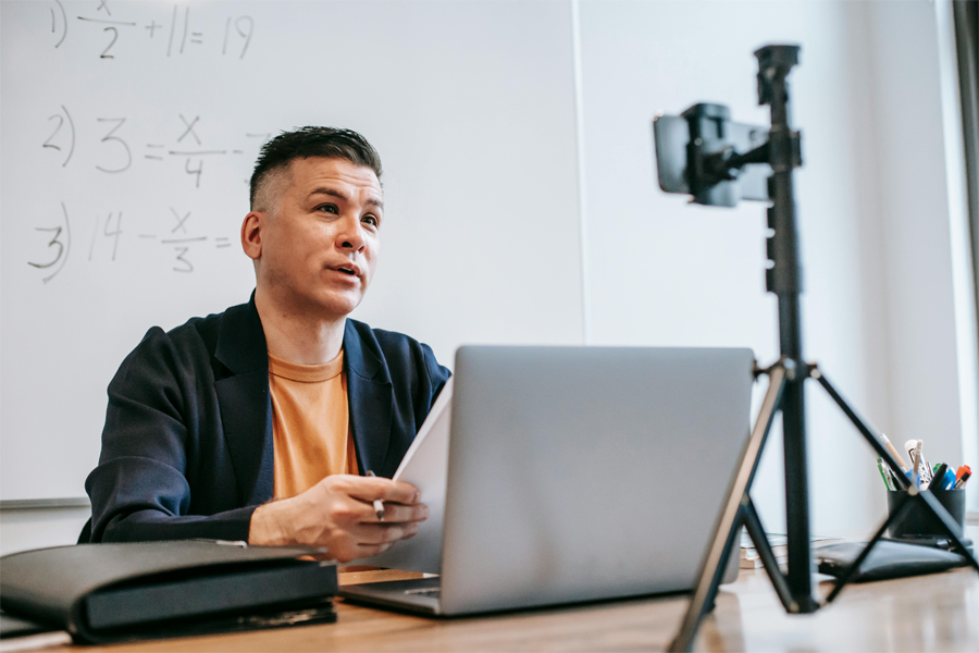 Male teacher sitting at a desk with papers in hand, looking at a smartphone camera set on a tripod in front of them.