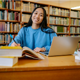 Older teenage girl sitting in a library smiling and in front of a laptop.
