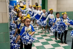 Young cheerleaders are lined up on either side of a school hallway chanting. They hold blue and white pom-poms and wear matching t-shirts and yellow bows in their hair.