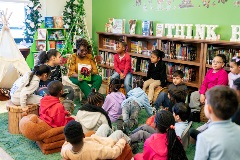 A group of students sitting on a rug in a green classroom library listen to a teacher who is reading a book aloud to them.