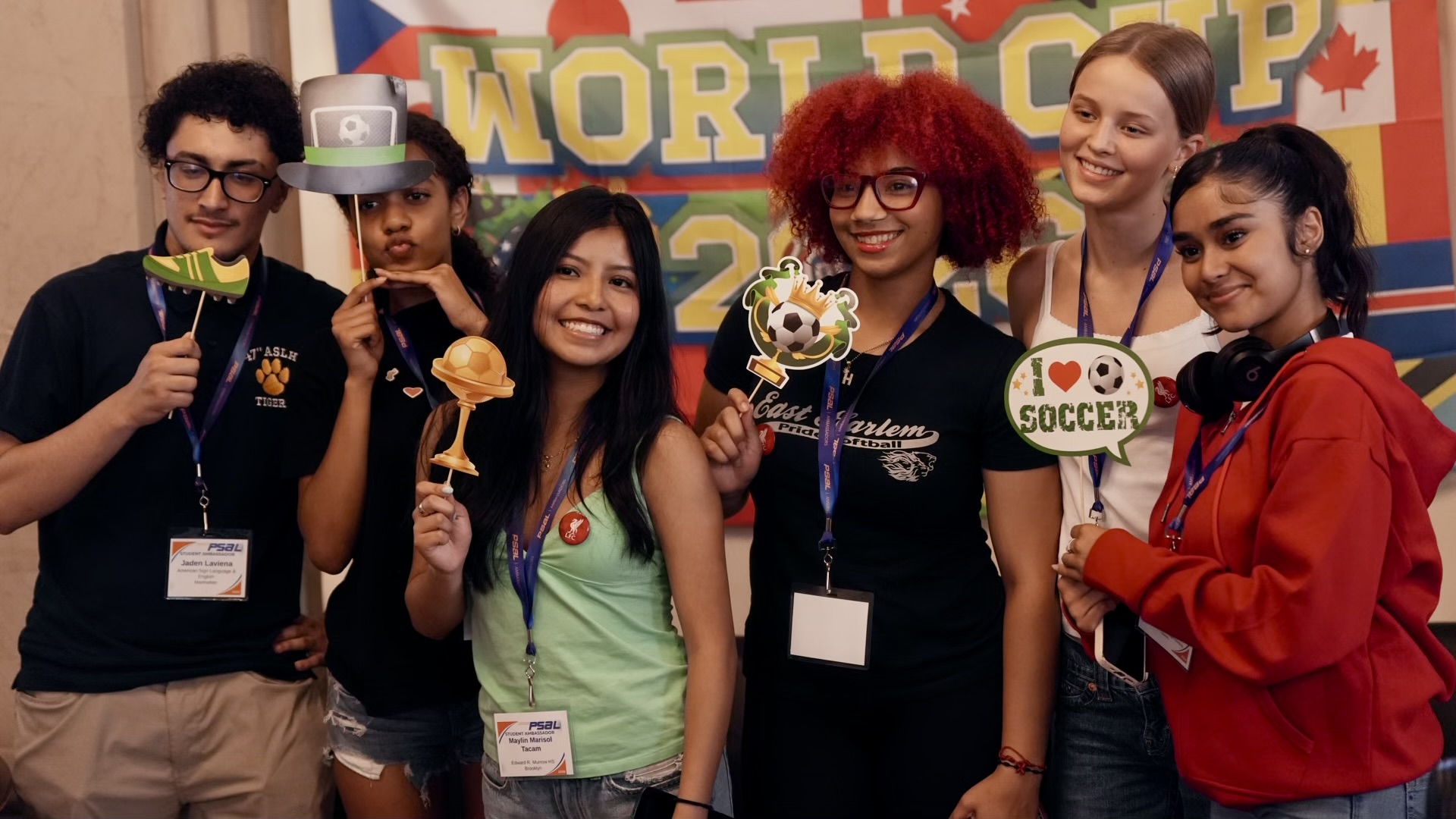 Group of six teenaged students holding soccer-related signs and decorations and standing in front of a banner that reads 'World Cup 2026.'