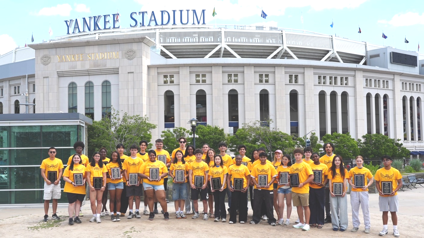 Group of over 30 high school-aged students holding award plaques and standing in front of Yankee Stadium