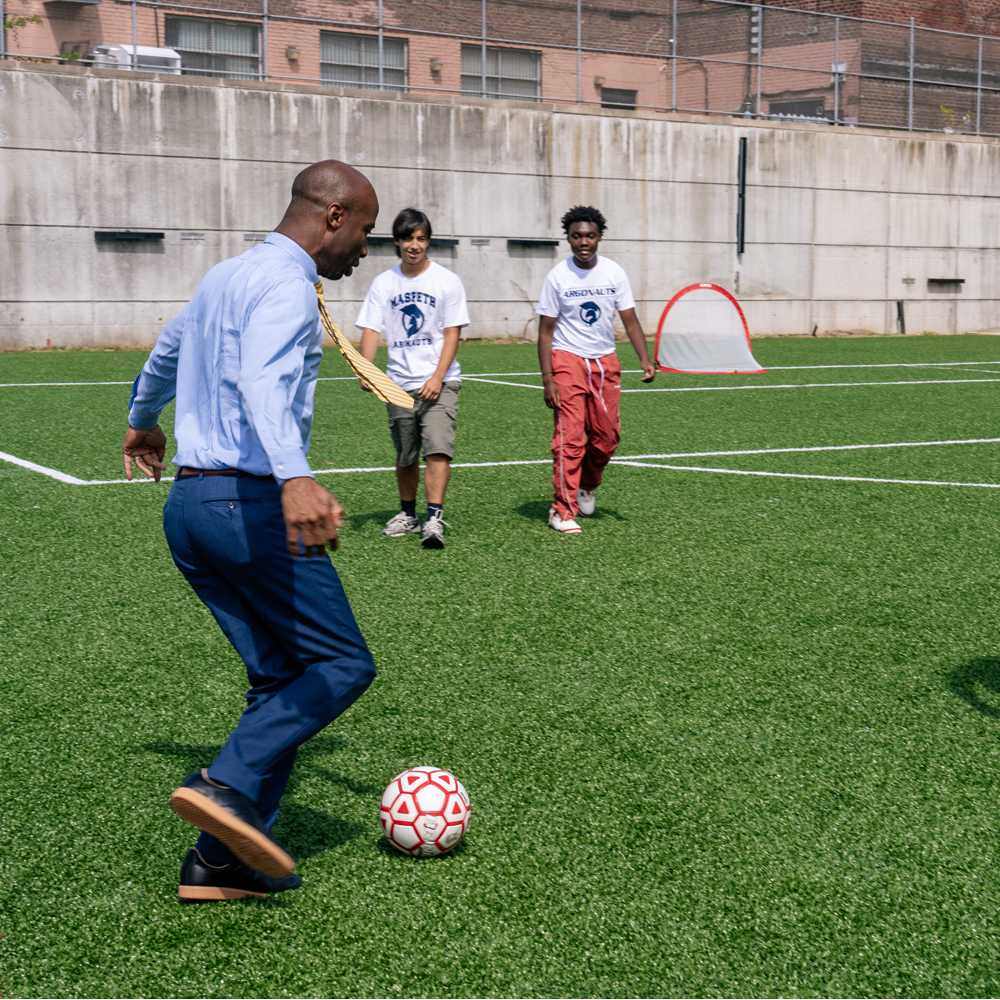 Chancellor Samuels getting ready to kick a soccer ball past two high school-aged students towards a goal.